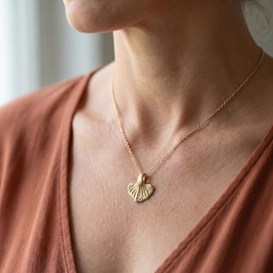 Puede incluir: Un collar dorado con un colgante en forma de abanico u hoja. El colgante cuelga de una delicada cadena. La persona lleva una blusa de color óxido. Joyería.