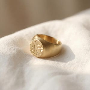 A gold ring with a decorative design sits on a white cloth.