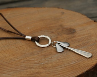 May include: A silver-toned pendant necklace with a hammered texture. The pendant features a long, slender shape with two smaller, leaf-like shapes attached. The necklace has a brown leather cord.