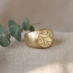 A gold ring with a leaf design sits on a beige fabric, accompanied by a sprig of greenery.