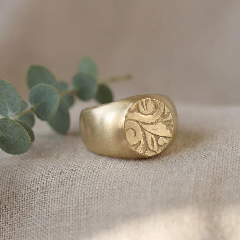 A gold ring with a leaf design sits on a beige fabric, accompanied by a sprig of greenery.