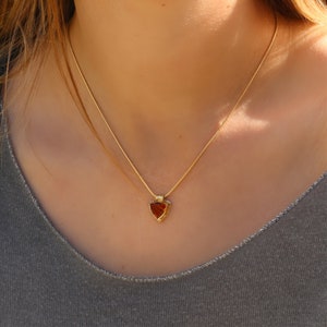 Puede incluir: Un collar de cadena de oro con un colgante triangular con una piedra roja.
