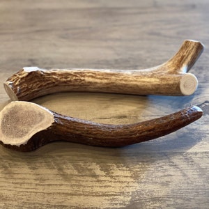 May include: Two natural deer antlers, one light brown and one dark brown, are displayed on a wooden surface. The antlers have a textured appearance and varying shades of brown and white. They are likely intended for pet use.
