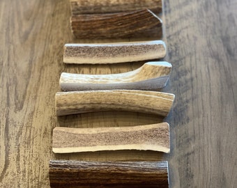 Puede incluir: Un conjunto de siete masticables de cuerno de ciervo de diferentes tamaños y formas. Los cuernos son naturales y tienen un color marrón claro. Los cuernos están dispuestos verticalmente sobre una superficie de madera con una regla en la parte inferior de la imagen.