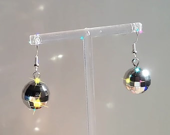 May include: A pair of silver disco ball earrings. Each earring features a small, spherical disco ball with reflective, mirrored facets. The earrings hang from silver-colored fish hook ear wires. The background is a light gray with sparkling highlights.