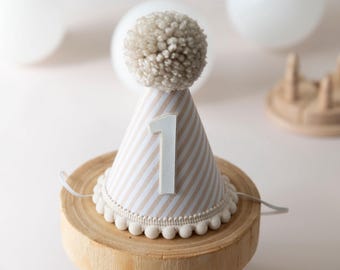 May include: A white and brown striped party hat with a white pom-pom on top and a white number "1" on the front. The hat is sitting on a wooden base.