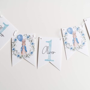 May include: A white banner with the name "Oliver" and the number "1" in light blue. Some banners feature a watercolour illustration of a rabbit holding a balloon, surrounded by a floral wreath. The banner is strung on a white string.