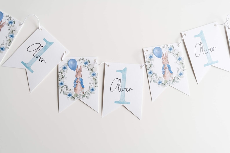 May include: A white banner with the name "Oliver" and the number "1" in light blue. Some banners feature a watercolour illustration of a rabbit holding a balloon, surrounded by a floral wreath. The banner is strung on a white string.