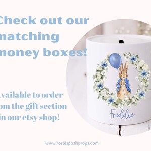 May include: White ceramic money box with a rabbit illustration holding a blue balloon, encircled by a floral wreath. The name "Freddie" is written below. Text on the image reads "Check out our matching money boxes!"