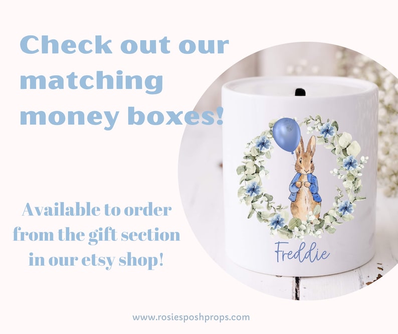 May include: White ceramic money box with a rabbit illustration holding a blue balloon, encircled by a floral wreath. The name "Freddie" is written below. Text on the image reads "Check out our matching money boxes!"