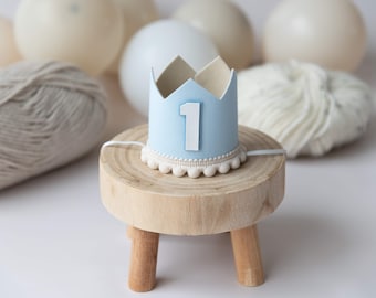 May include: A light blue paper crown with a white number one on it. The crown is sitting on a small wooden stool.