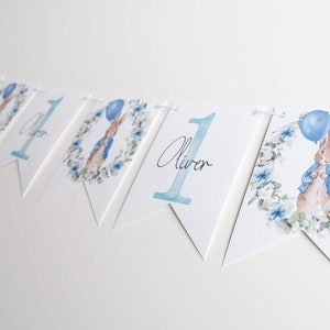 May include: A white bunting with blue watercolour illustrations. The bunting features the number "1", the name "Oliver", and images of a rabbit holding a blue balloon. The rabbit is surrounded by a floral wreath.