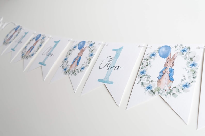 May include: A white bunting with blue watercolour illustrations. The bunting features the number "1", the name "Oliver", and images of a rabbit holding a blue balloon. The rabbit is surrounded by a floral wreath.