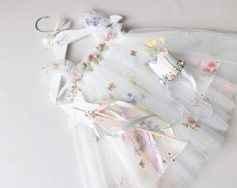 May include: A white tulle dress with floral and butterfly accents, displayed on a white hanger with the text "Rosie's Boutique Design". A star-shaped wand and a crown with floral details complete the fairy-themed ensemble.