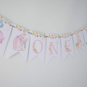 May include: A white and pink banner with the word "ONE" in pink letters. The banner is decorated with floral designs and images of Peter Rabbit characters.