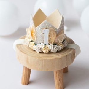 May include: A white felt crown with a glittery centre and a number one on top. The crown is decorated with white and yellow flowers and two butterfly wings. The crown is sitting on a small wooden stool.