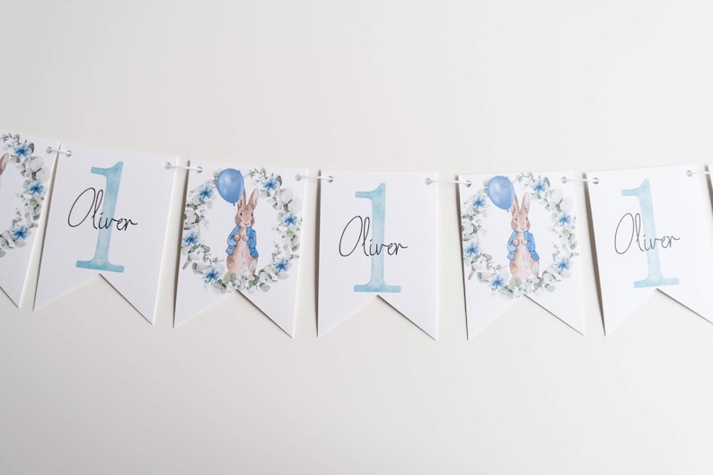 May include: A decorative banner with white triangular flags. Each flag displays the number 1 in pale blue and the name "Oliver". Some flags feature a Peter Rabbit illustration with a blue balloon and floral wreath.
