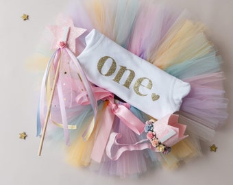 Peut inclure: Une jupe tutu en tulle arc-en-ciel, un body blanc avec le texte "one" en paillettes dorées, une baguette rose avec un sommet en étoile et une couronne rose avec des fleurs et un "one" blanc dessus.