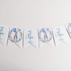 May include: A bunting banner with white pennants. Each pennant displays a blue number 1, the name "Oliver", and a Peter Rabbit illustration with a balloon. The design includes a floral wreath, ideal for a first birthday.