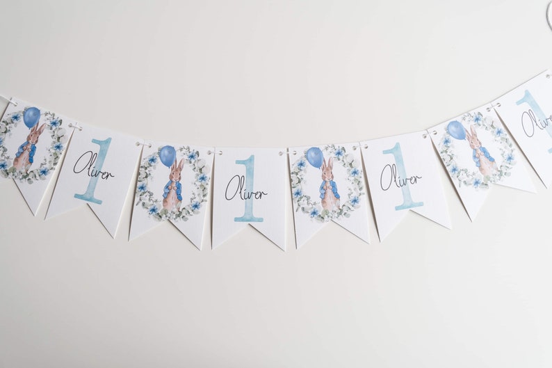 May include: A bunting banner with white pennants. Each pennant displays a blue number 1, the name "Oliver", and a Peter Rabbit illustration with a balloon. The design includes a floral wreath, ideal for a first birthday.