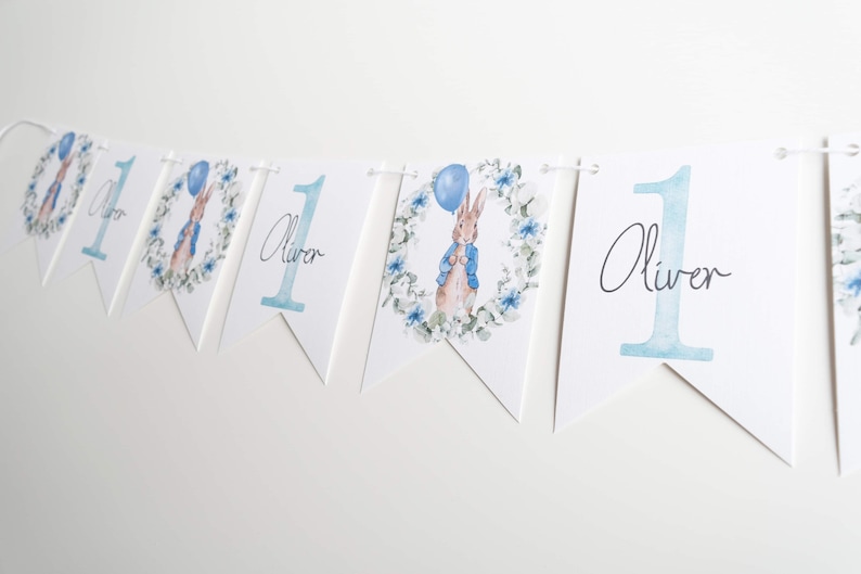 May include: A white bunting banner with blue details. It features a watercolour illustration of Peter Rabbit with a blue balloon, framed by a floral wreath. The banner also displays the number "1" and the name "Oliver". Ideal for a first birthday celebration.