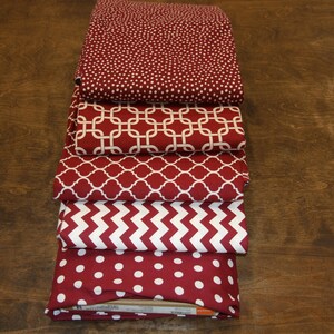 May include: A stack of folded fabric pieces in various patterns. The fabrics are primarily burgundy with white designs, including polka dots, geometric shapes, and chevrons. The fabrics are stacked on a dark wooden surface.