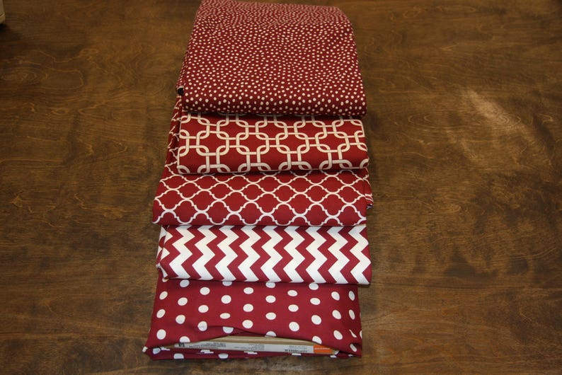 May include: A stack of folded fabric pieces in various patterns. The fabrics are primarily burgundy with white designs, including polka dots, geometric shapes, and chevrons. The fabrics are stacked on a dark wooden surface.
