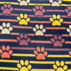 May include: Navy blue fleece fabric featuring a repeating pattern of paw prints and stripes. The paw prints are in yellow, orange, cream, and burgundy. The stripes are yellow and orange, creating a vibrant design.