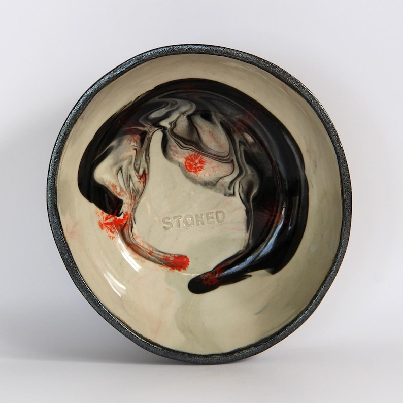 Puede incluir: Un cuenco de cer&aacute;mica con interior crema y borde gris oscuro. El cuenco presenta un dise&ntilde;o abstracto en negro y rojo con la palabra "STOKED" en relieve en el interior. El cuenco es redondo y tiene una forma ligeramente irregular.