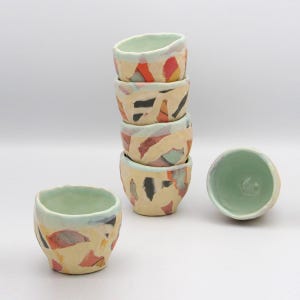 May include: A stack of five small ceramic bowls with a light green interior and a beige exterior decorated with colorful abstract shapes. One bowl is separate, and another is turned upside down.