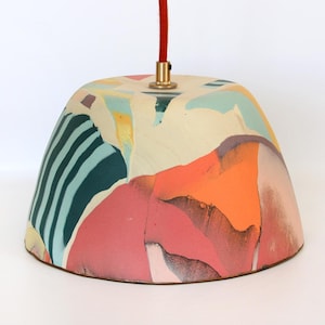 May include: A dome-shaped pendant light shade featuring an abstract pattern of teal, orange, pink, and cream colours. The shade is suspended by a red fabric cord attached to a brass-coloured metal fixture at the top centre of the dome.