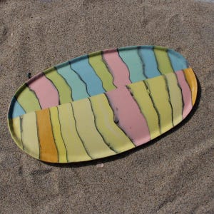 May include: An oval ceramic serving platter with a colorful, abstract design. The platter is divided into two sections, each with vertical stripes in pastel shades of yellow, pink, blue, and green, separated by black lines. The platter is resting on sand.