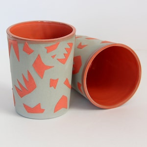May include: Two ceramic mugs with a geometric pattern. The mugs are gray with a bright orange geometric pattern. The inside of the mugs is orange.