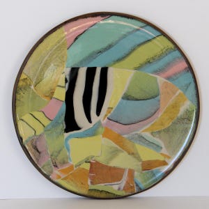 May include: A round decorative plate with a colorful abstract design. The plate features a mosaic of shapes in shades of yellow, green, blue, pink, and orange, with a black and white striped section. The plate has a dark brown rim.