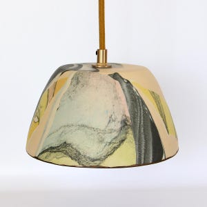 May include: A pendant light fixture with a beige, bell-shaped shade. The shade features a colorful abstract design with yellow, black, and gray elements. A gold-colored metal fixture connects the shade to a gold-colored cord.