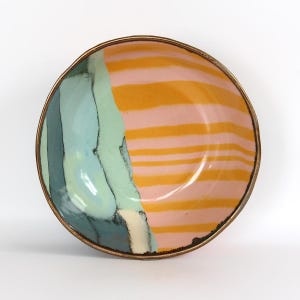 May include: A round ceramic bowl with a dark metallic rim. The interior features a split design: one side displays abstract, flowing shapes in shades of teal, light blue, and cream, while the other side shows horizontal orange stripes on a pink background.