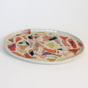 Może przedstawiać: Owalny ceramiczny półmisek do serwowania z mozaikowym wzorem. Półmisek ma jasnoszarą krawędź i powierzchnię ozdobioną kolorowymi, nieregularnymi kształtami w odcieniach czerwieni, pomarańczy, różu, zieleni i czerni. Ogólna estetyka jest artystyczna i nowoczesna.