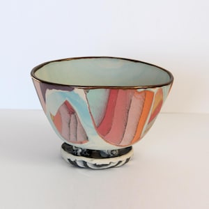 May include: A colorful ceramic bowl with a light blue interior and a decorative exterior featuring vertical stripes in shades of red, orange, and blue. The bowl has a dark rim and sits on a patterned base.