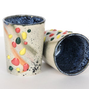 May include: Two ceramic mugs with a speckled glaze and colorful dots. The mugs are a light beige color with a dark blue rim and interior.