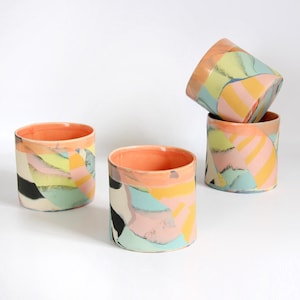 May include: Three cylindrical ceramic cups featuring a vibrant abstract pattern of pastel pink, yellow, light blue, and black shapes. The interior of each cup is finished in a solid, smooth peach-orange glaze, contrasting with the colorful exterior.