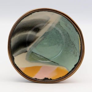 May include: A small, round ceramic bowl with a bronze-colored rim. The interior features a colorful abstract design with shades of black, green, yellow, and pink. The bowl's design is a mix of brushstrokes and color blocks.