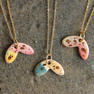 Puede incluir: Tres collares de cadena dorada con colgantes. Los colgantes tienen forma de bumerán, con un diseño moteado en rosa, amarillo y azul. Los colgantes tienen un lazo de metal dorado para la cadena.