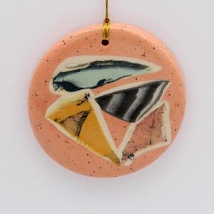 May include: A round, pink ceramic ornament with a speckled texture. The ornament features a design of abstract shapes in shades of yellow, blue, black, and white.