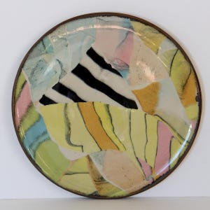 May include: A round ceramic plate with a brown rim. The plate's surface is a collage of colorful abstract shapes in yellow, pink, blue, and orange, with a central black and white striped design. The plate has a glossy finish.