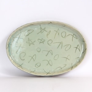 May include: Oval ceramic serving platter with a light green glaze. The platter is decorated with hand-drawn gray symbols and letters. The rim has a slightly textured, uneven edge. The overall design is simple and artistic.