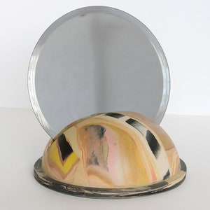Peut inclure: Un dôme rond en céramique avec un motif tourbillonnant de noir, blanc, jaune, rose et marron. Un miroir rond argenté est posé sur le dôme.