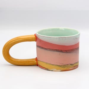 May include: A handmade ceramic mug with a large, curved handle. The mug features a colorful design with horizontal bands of pink, white, red, yellow, and black. The interior is a light green color. The handle is a mustard yellow color.