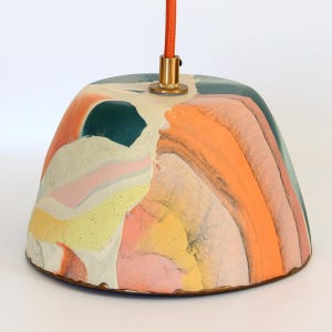 May include: A dome-shaped pendant light fixture featuring a marbled pattern of orange, pink, yellow, teal, and white colors. The lamp has a metallic gold-toned top fitting and is suspended by a bright orange fabric-covered electrical cord.