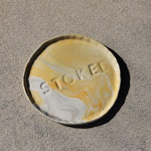 Può includere: Un piatto rotondo in ceramica con un design marmorizzato giallo, bianco e grigio. La parola "STOKED" è in rilievo al centro. Il piatto ha un bordo leggermente ondulato ed è posto su uno sfondo neutro.