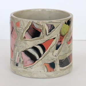 May include: A cylindrical ceramic planter with a white glaze and a mosaic design. The mosaic is made of small, irregular pieces of ceramic in shades of pink, orange, black, and white.